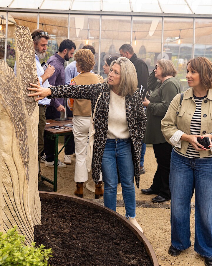 A woman reaches out to touch one of Richard Clegg's structure, whilst another woman looks on. There are people stood behind them in front of The Glasshouse.